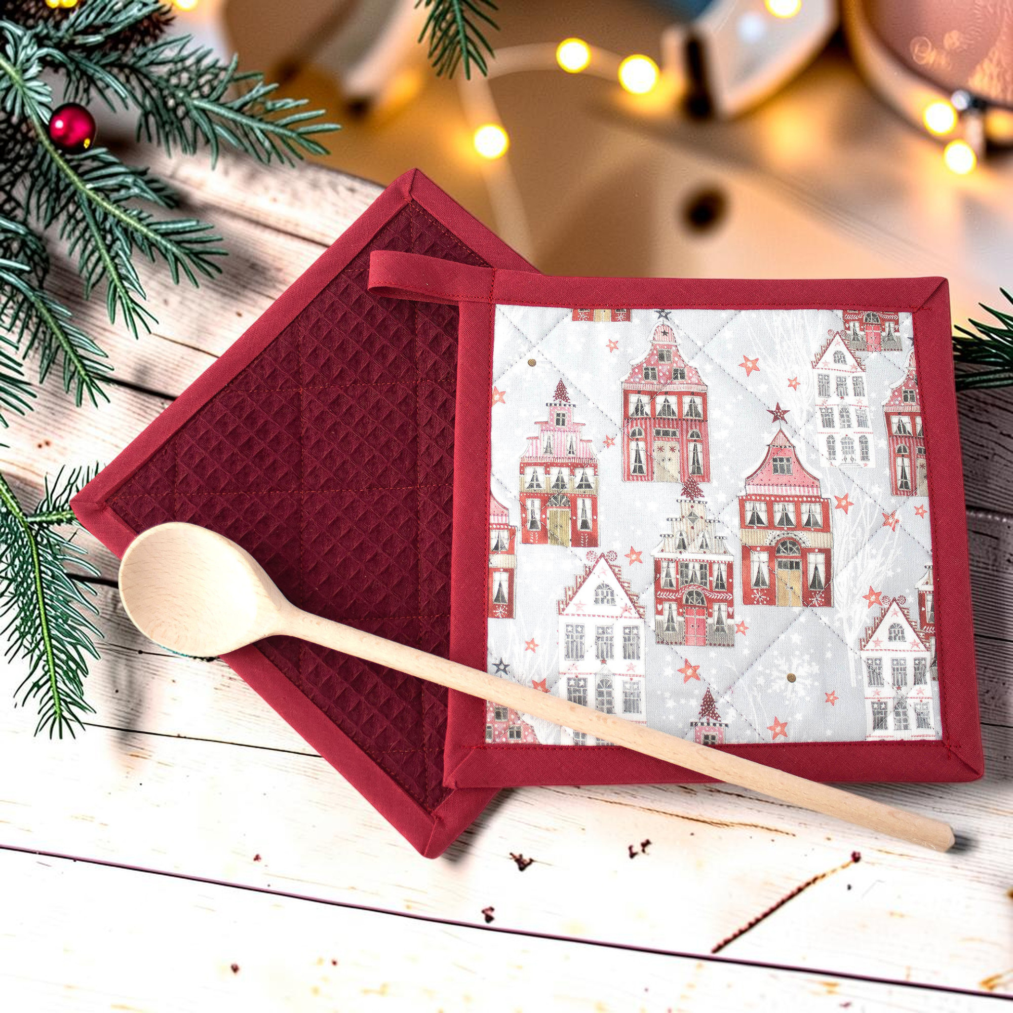 Weihnachtliche Topflappen mit Häusern und Sternen, dekorativ mit Holzlöffel und Tannenzweigen vor festlicher Kulisse