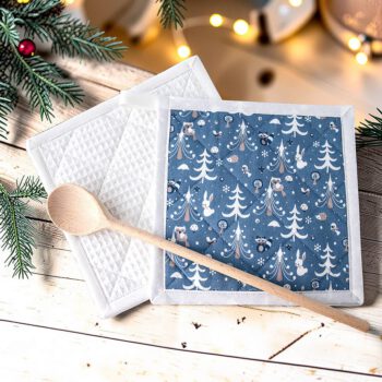 Handgefertigte Topflappen mit blauem Winterwald-Motiv, weißen Bäumen, Tieren und Pilzen, auf einem Holztisch mit Weihnachtsdekoration